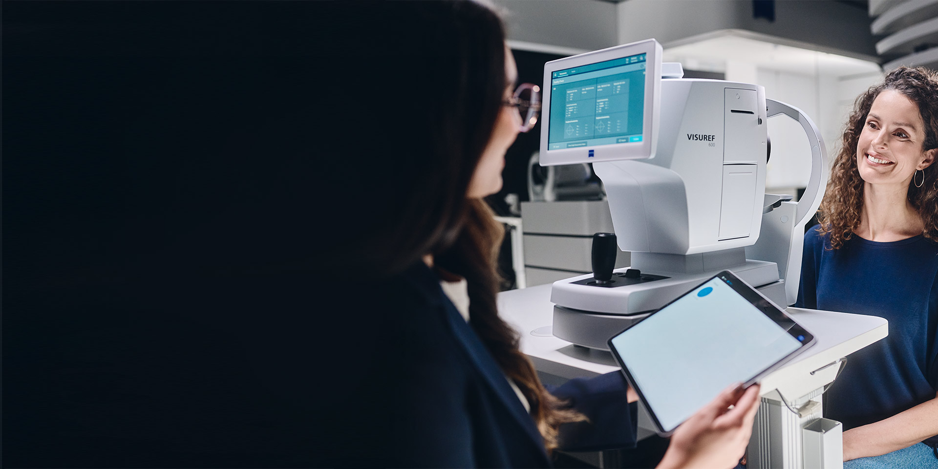 Жінка-оптик дивиться на свій планшет, сидячи перед приладом ZEISS VISUREF 600 та розмовляючи зі своїм пацієнтом.