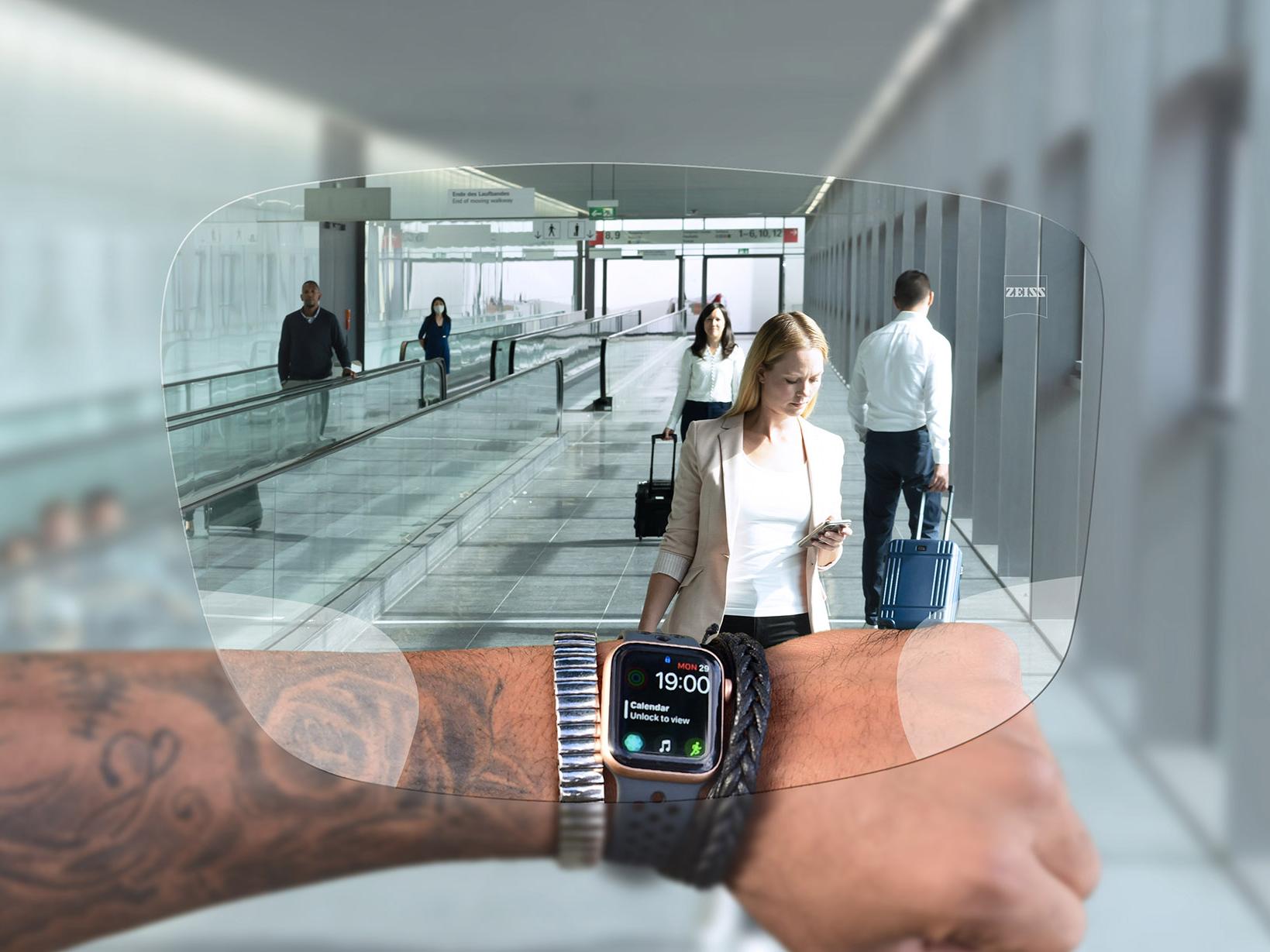 Людина дивиться на годинник крізь лінзи ZEISS SmartLife Digital. На тлі показаний типовий аеропорт – люди йдуть по ескалаторах і везуть із собою валізи. 
