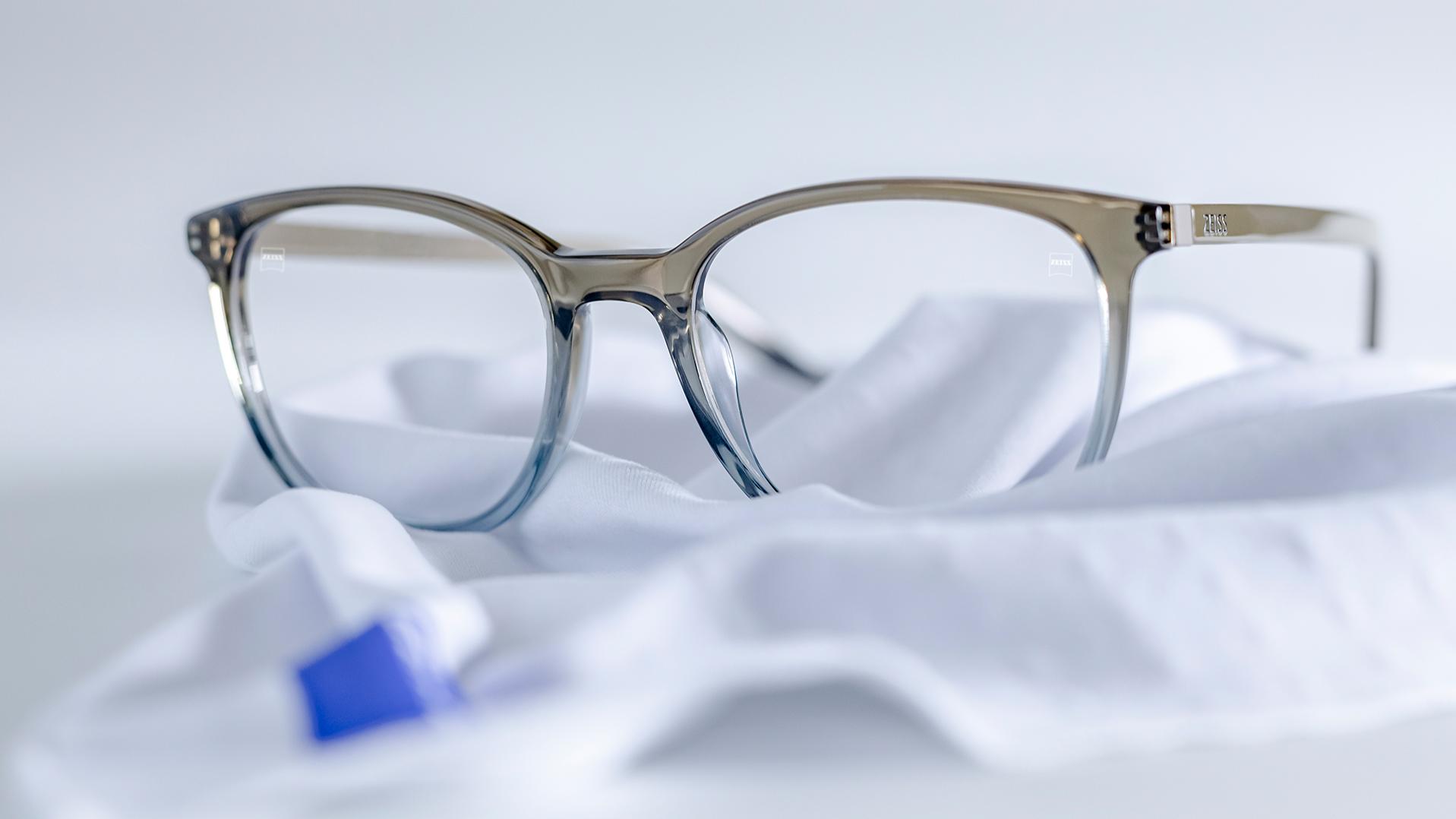 Окуляри з сіро-блакитною оправою та лінзами ZEISS із покриттям DuraVision® лежать на білій серветці з мікроволокна.