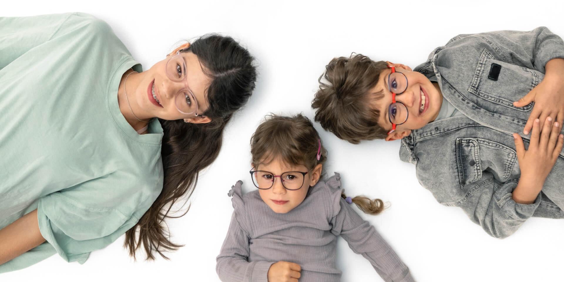 Підліток і двоє молодших дітей в окулярах із лінзами ZEISS Myopia лежать на підлозі.
