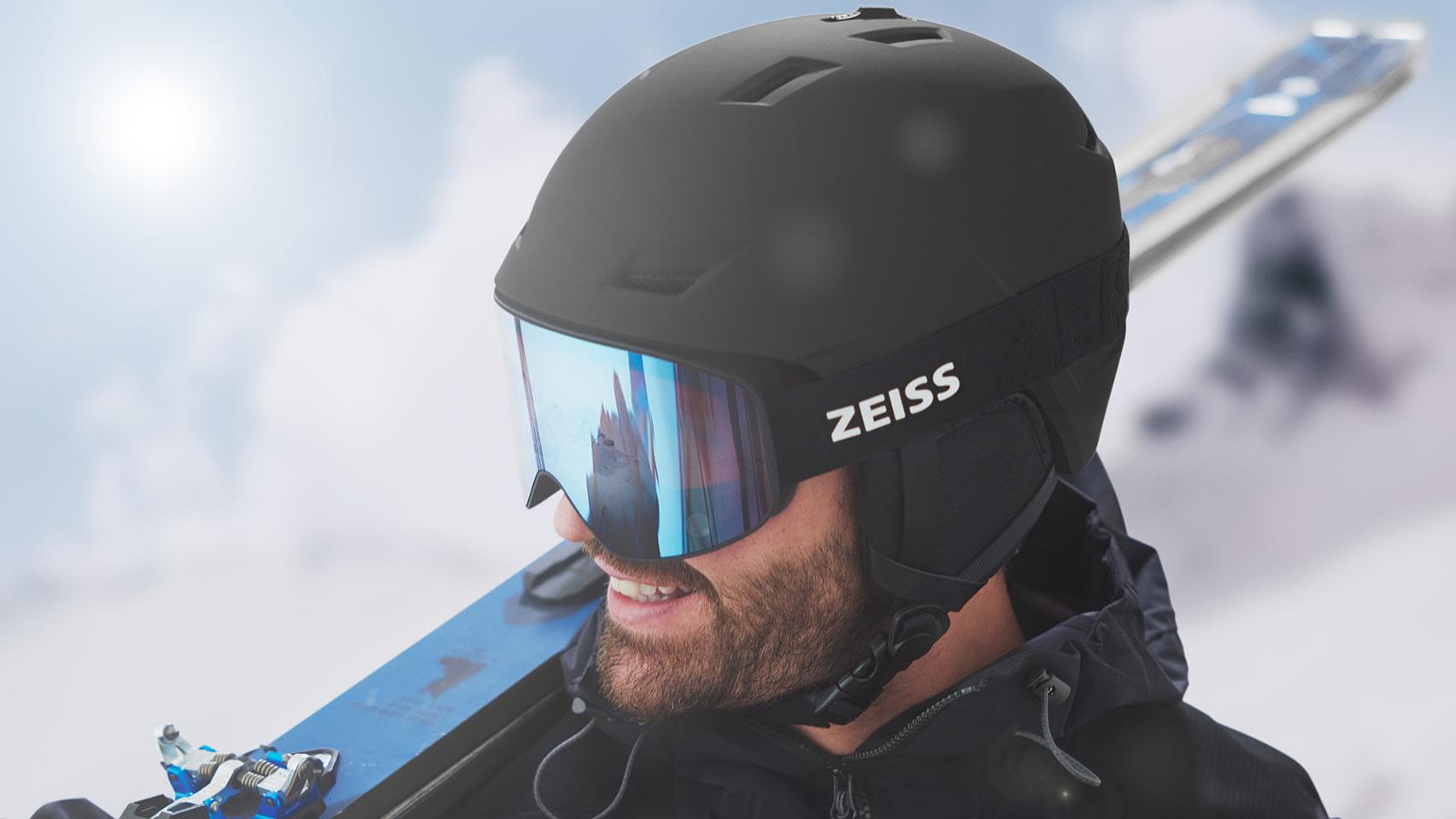 Чоловік у чорному шоломі й синій гірськолижній масці ZEISS із дзеркальним ефектом тримає лижі на правому плечі.